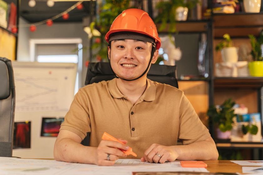 Asian male engineer wearing safety helmet, smiling at desk in office environment.