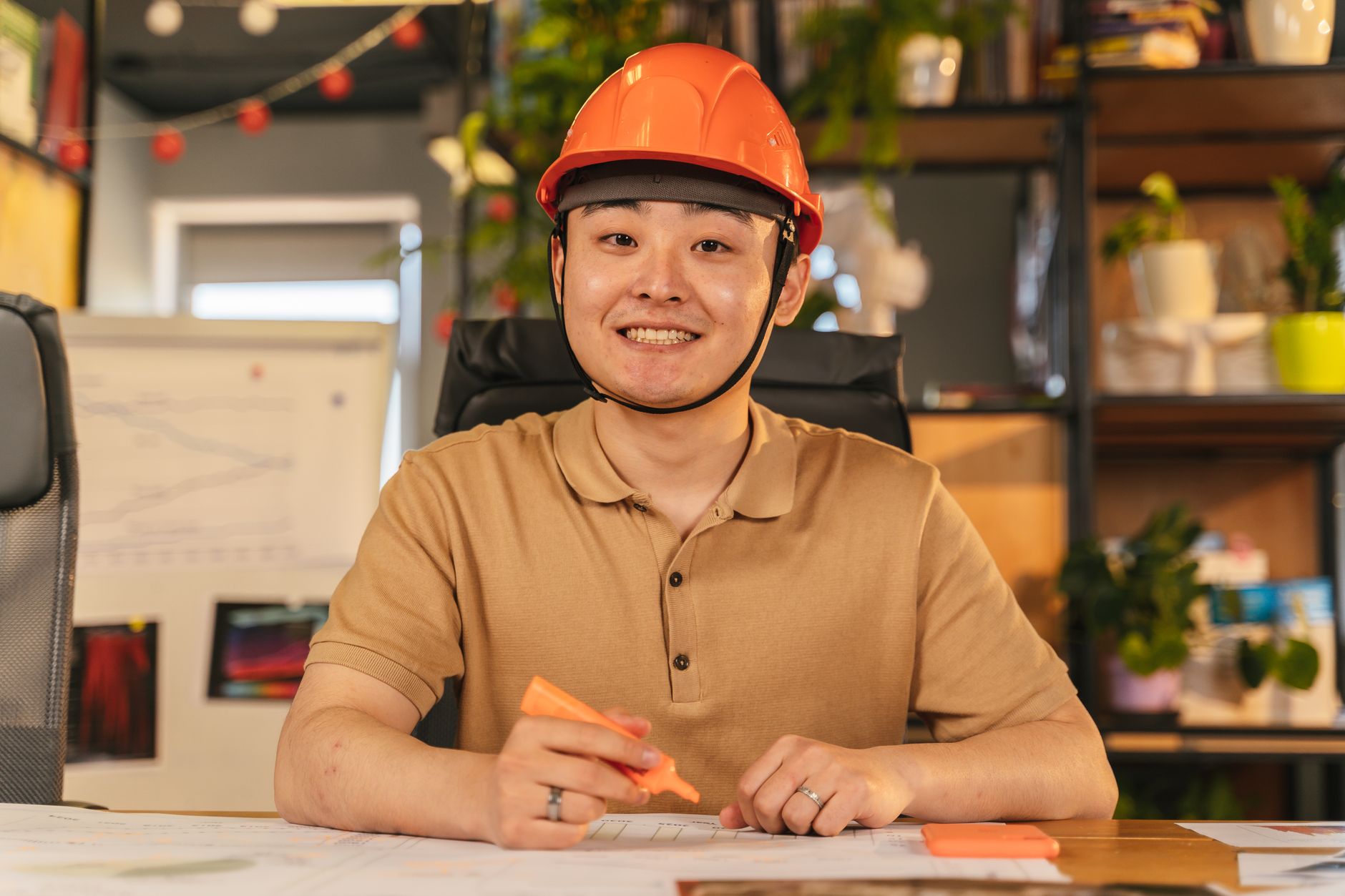 Asian male engineer wearing safety helmet, smiling at desk in office environment.